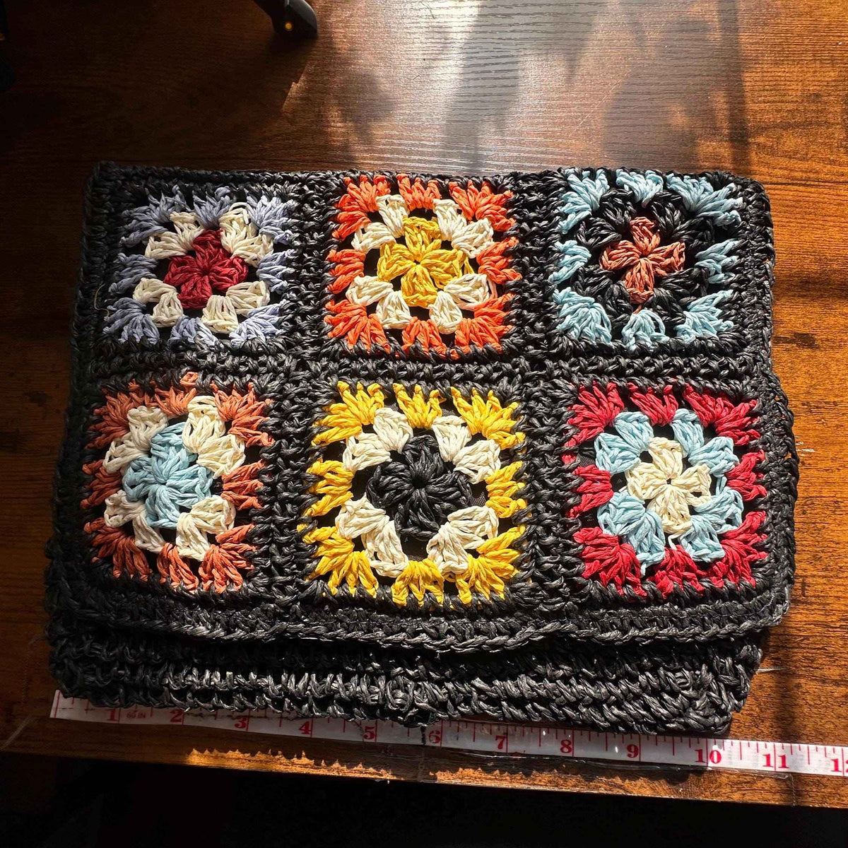 Handmade crochet granny square purse featuring colorful floral patchwork and textured black crochet trim with dimension ruler displayed on table