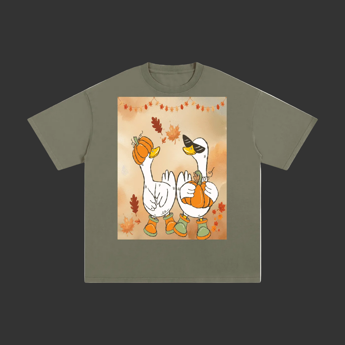 An olive green short-sleeve t-shirt with a central graphic print of two white geese wearing orange pumpkins and boots, surrounded by autumn leaves, shown on a plain gray background.