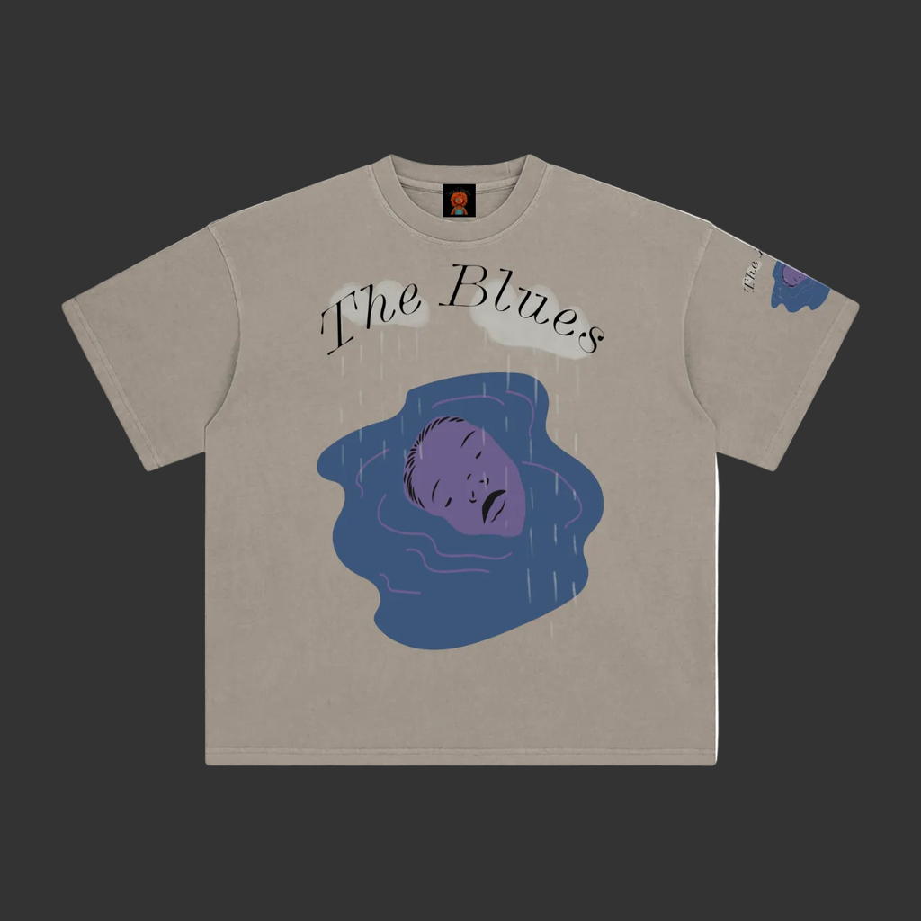 A beige crewneck t-shirt featuring a graphic print of a purple face in blue water with the text 'The Blues' written above, displayed flat against a gray background.
