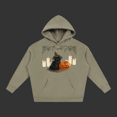 A sage green pullover hoodie with a Halloween graphic on the chest, featuring a black cat in a witch hat next to a jack-o'-lantern pumpkin and white candles, shown on a plain gray background.
