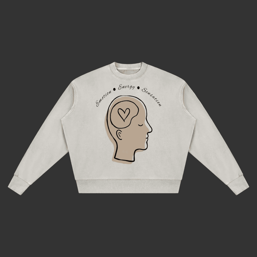 A light beige crewneck sweatshirt featuring a graphic of a side-profile human head with a heart inside the brain, with the words 'Sensation • Energy • Simulation' written above, shown on a plain gray background.