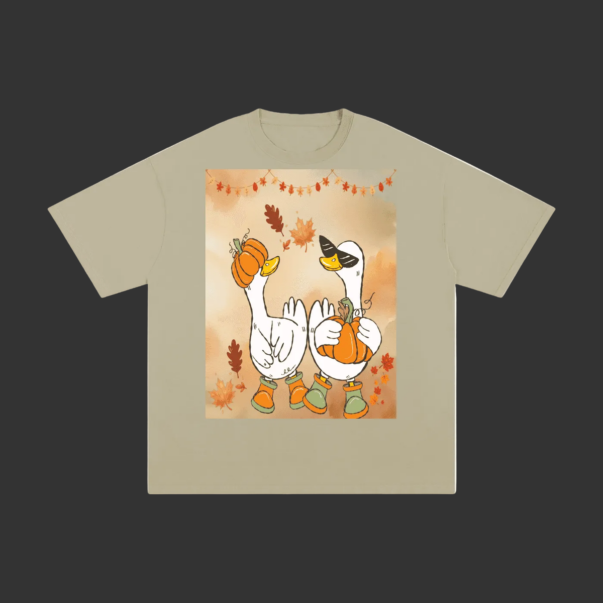 A beige short-sleeve t-shirt with a central graphic print of two white geese wearing orange pumpkins and boots, set against a warm autumn background with leaves and a string of lights.