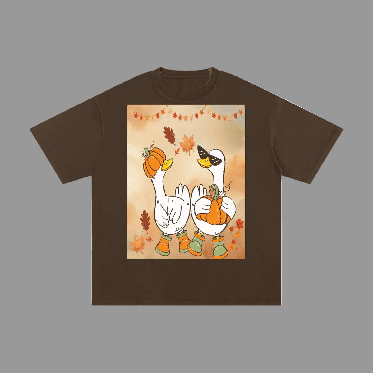 A brown short-sleeve t-shirt with a central graphic print of two white geese wearing black sunglasses and holding orange pumpkins, surrounded by autumn leaves and a string of lights.