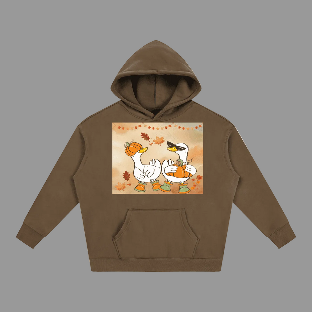 A brown pullover hoodie with a front kangaroo pocket and a central square graphic showing two cartoon swans holding pumpkins under a warm autumn sky with falling leaves.