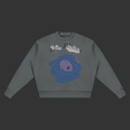 A dark gray crewneck sweatshirt featuring a graphic print of a purple figure in a blue puddle under gray clouds with the text 'The Blues' written above, displayed on a solid gray background.