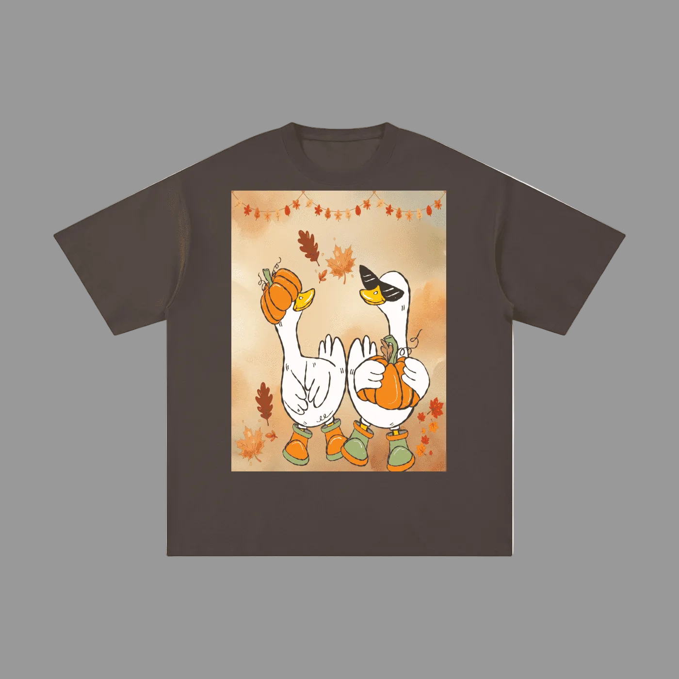 A dark brown short-sleeve t-shirt with a large square graphic print on the front. The graphic features a cartoon illustration of two white geese wearing orange pumpkins and green boots, surrounded by autumn leaves and a string of lights, set against a warm beige background.
