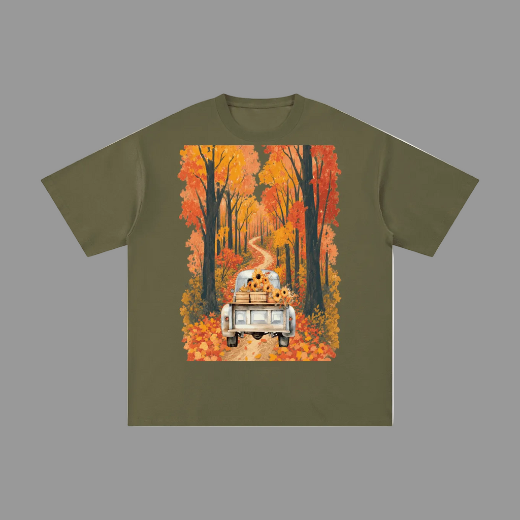 An olive green short-sleeve t-shirt with a large square graphic print on the front. The graphic shows a white pickup truck driving down a winding road through a forest of orange and red autumn trees, with sunflowers in the back of the truck.