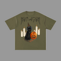 An olive green short-sleeve t-shirt with a Halloween graphic print featuring a black cat in a witch hat, a jack-o'-lantern pumpkin, and white candles, displayed on a solid grey background.
