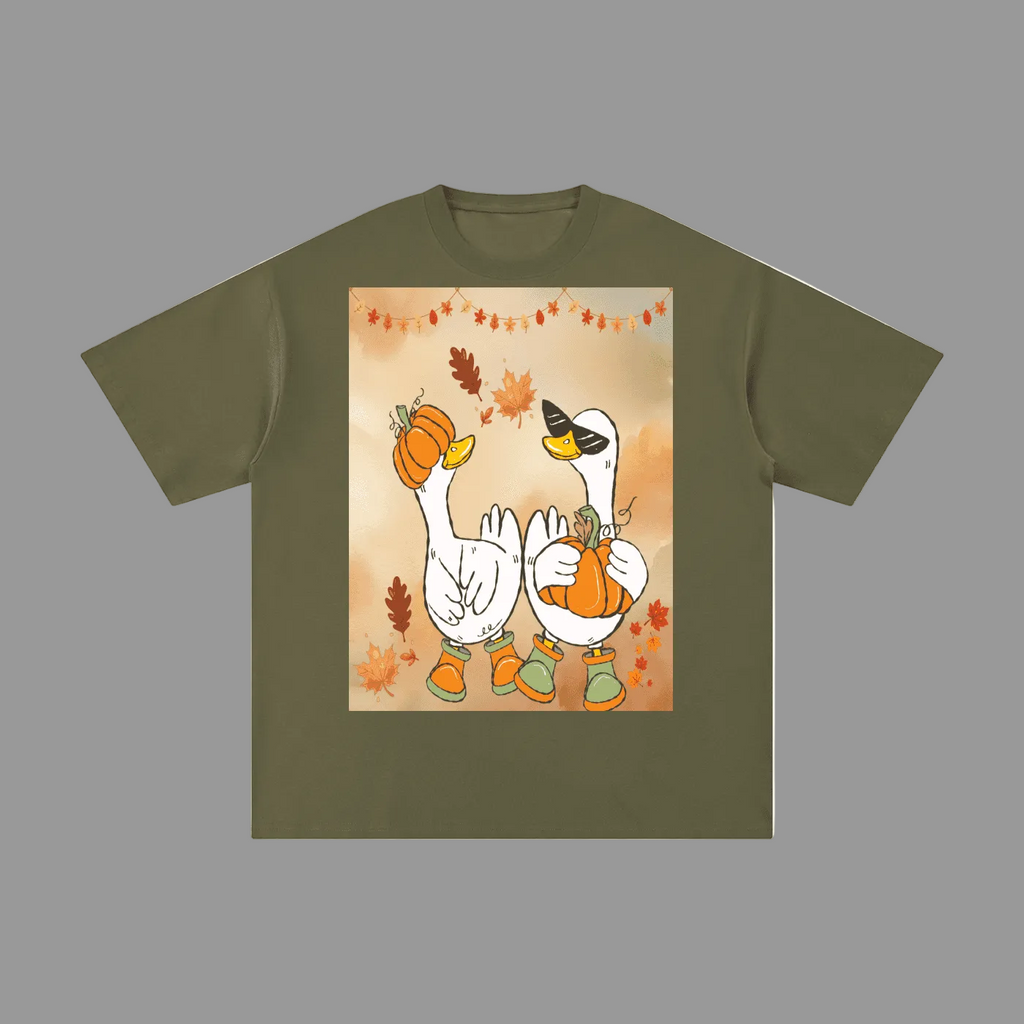 An olive green short-sleeve t-shirt with a central square graphic print of two cartoon white geese wearing orange boots and holding pumpkins, surrounded by autumn leaves on a warm beige background.