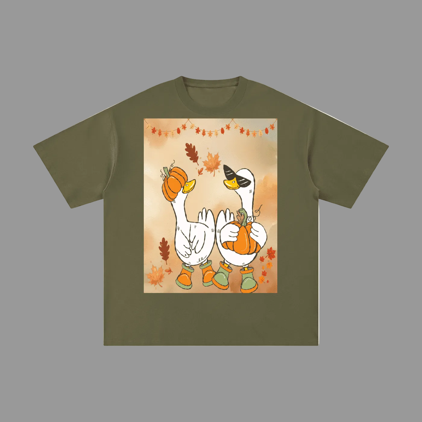 An olive green short-sleeve t-shirt with a central square graphic print of two cartoon white geese wearing orange boots and holding pumpkins, surrounded by autumn leaves on a warm beige background.