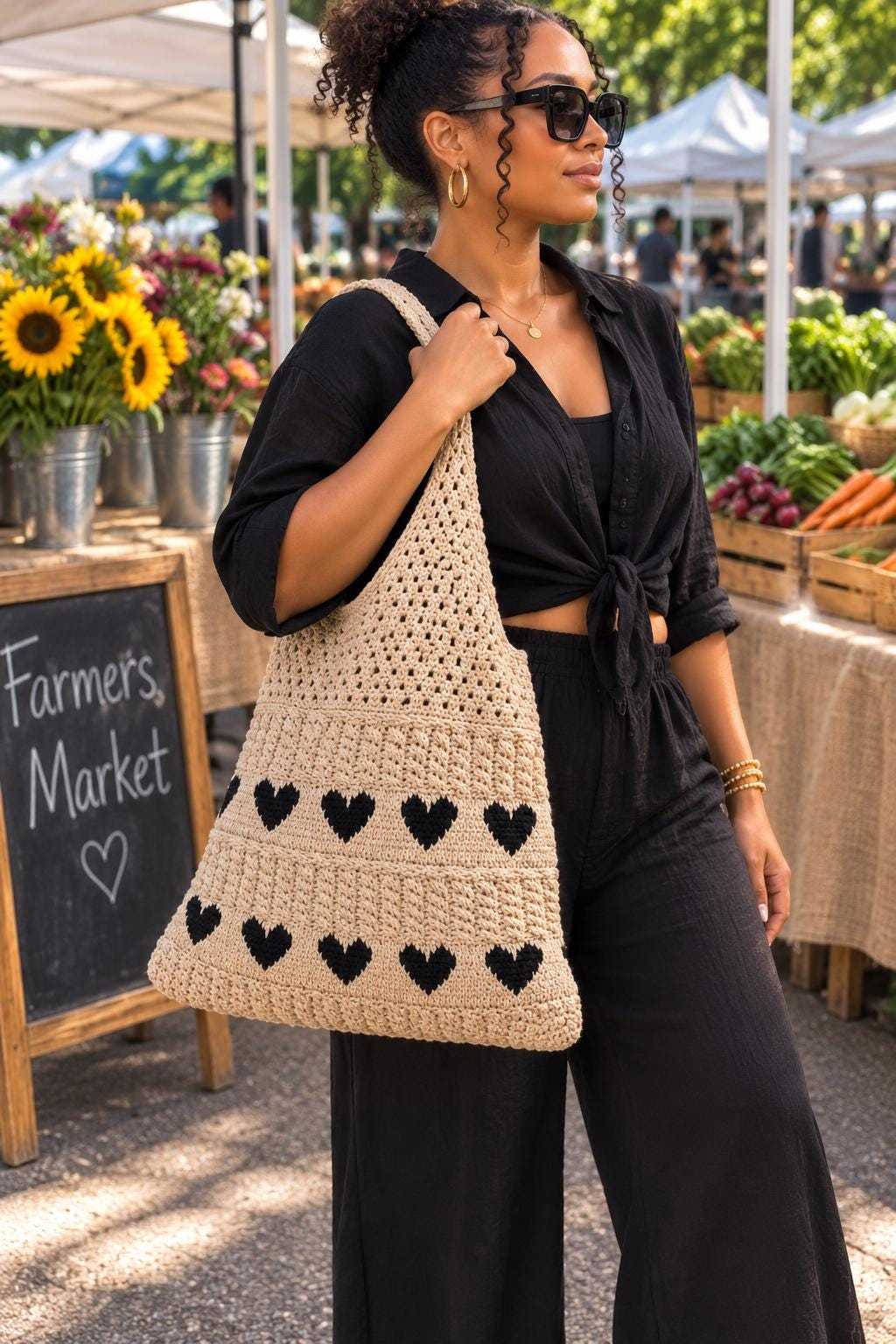 A woman at an outdoor farmers market wearing a black outfit and sunglasses, carrying a large beige crocheted shoulder bag with black heart patterns.