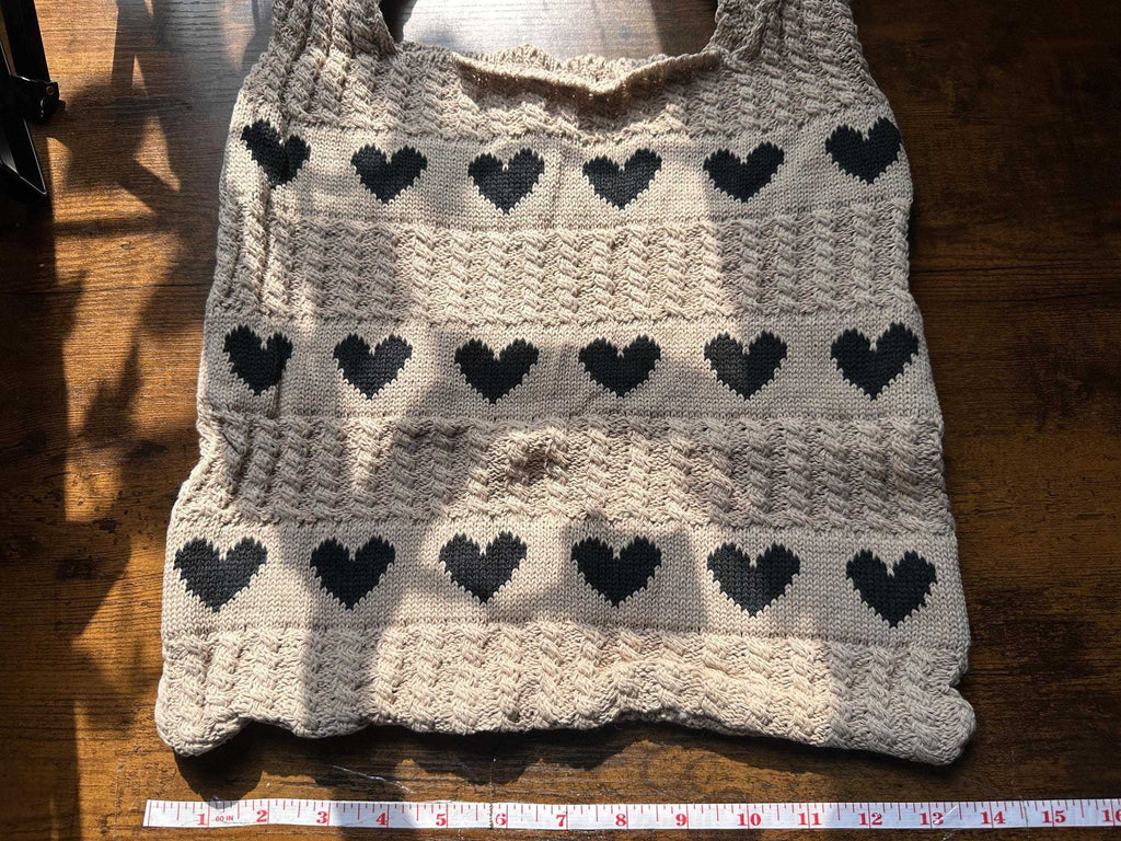 A cream-colored knit sweater vest with a black heart pattern, laid flat on a wooden surface with a measuring tape visible at the bottom.