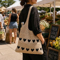 A woman at an outdoor farmers market wearing a large, chunky cream-colored crochet shoulder bag with black heart patterns. The background shows market stalls, other shoppers, and chalkboard signs