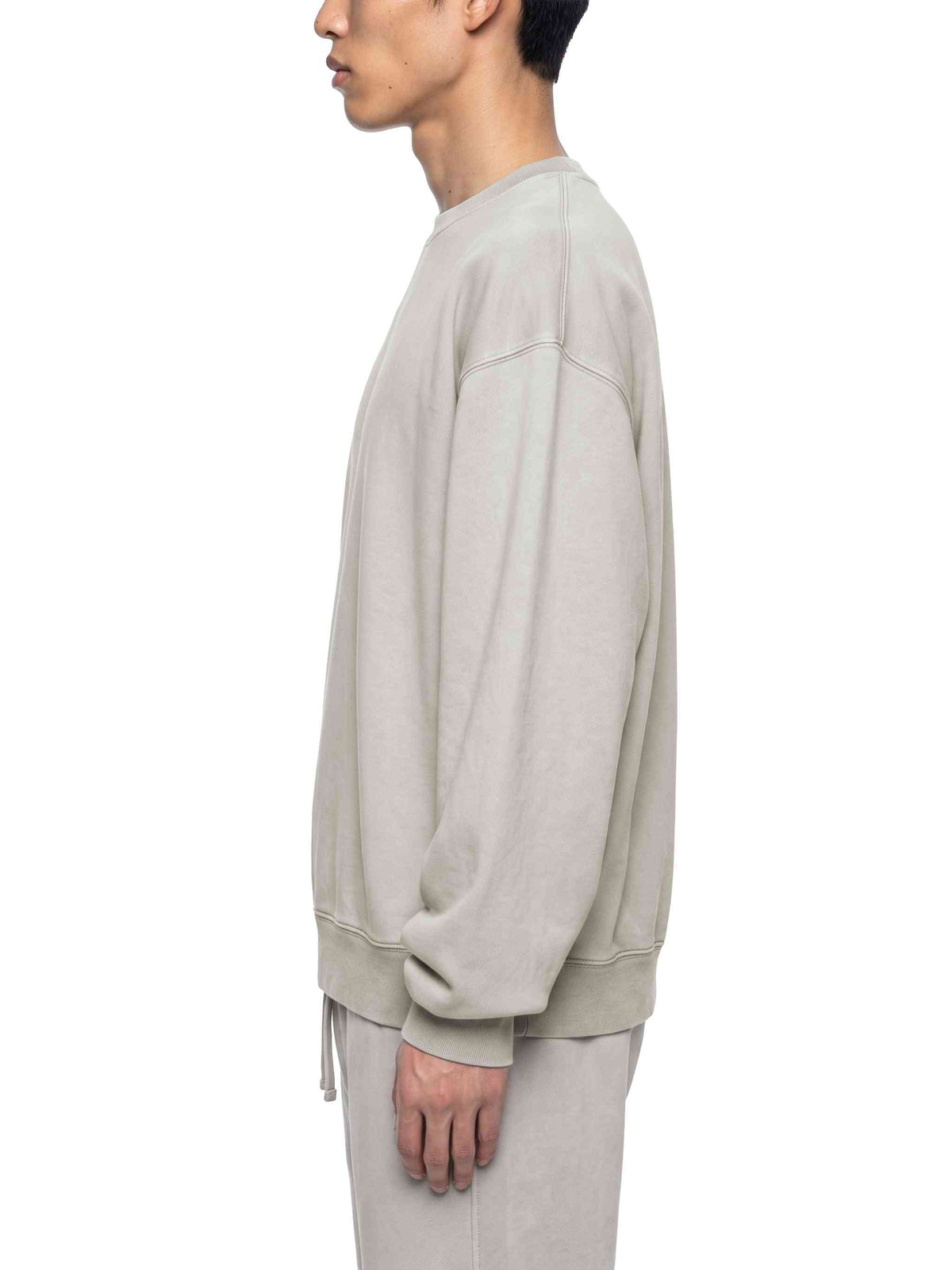 A man shown in profile wearing an oversized light beige crewneck sweatshirt and matching sweatpants against a plain white background.